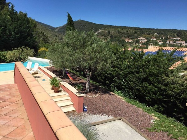 A pool area with steps leading down to a garden, surrounded by trees and a view of a hillside with buildings.