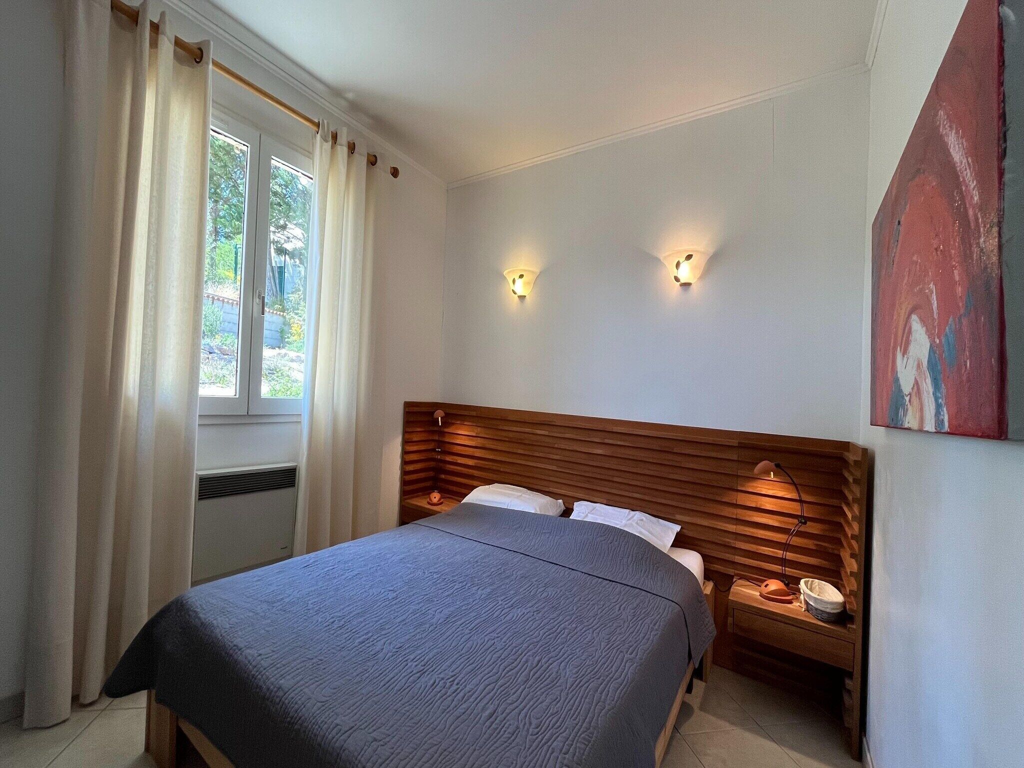 A bedroom with a bed, nightstands, a window with curtains, and wall-mounted lights.