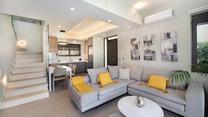Una sala de estar moderna con un sofá gris, cojines amarillos y una mesa de centro blanca. Una escalera conduce a un nivel superior, y se ve un área de cocina al fondo.