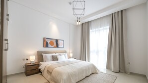 Ein modernes Schlafzimmer mit einem großen Bett, Nachttischen, einem an der Wand befestigten Kunstwerk und Blick ins Freie durch transparente Vorhänge.