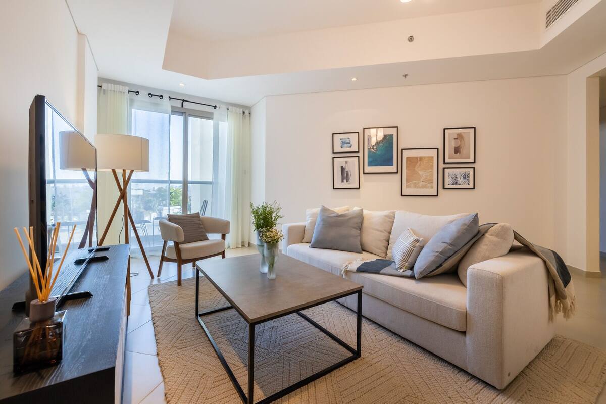 Una sala de estar moderna con sofá, mesa de centro y un espejo. Hay cuadros enmarcados en la pared y una ventana con cortinas.