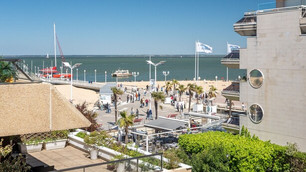 Un paseo marítimo frente a la playa, con un muelle para barcos y un edificio moderno con un ventanal circular.