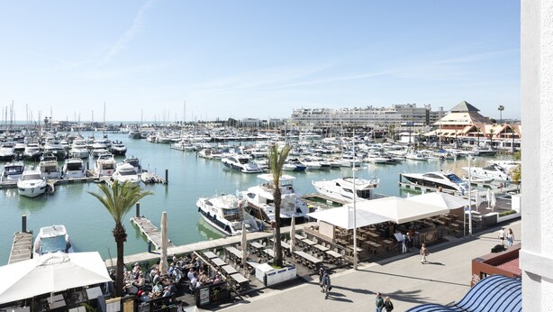 A marina with numerous boats docked, a waterfront restaurant area, and a large building in the background.