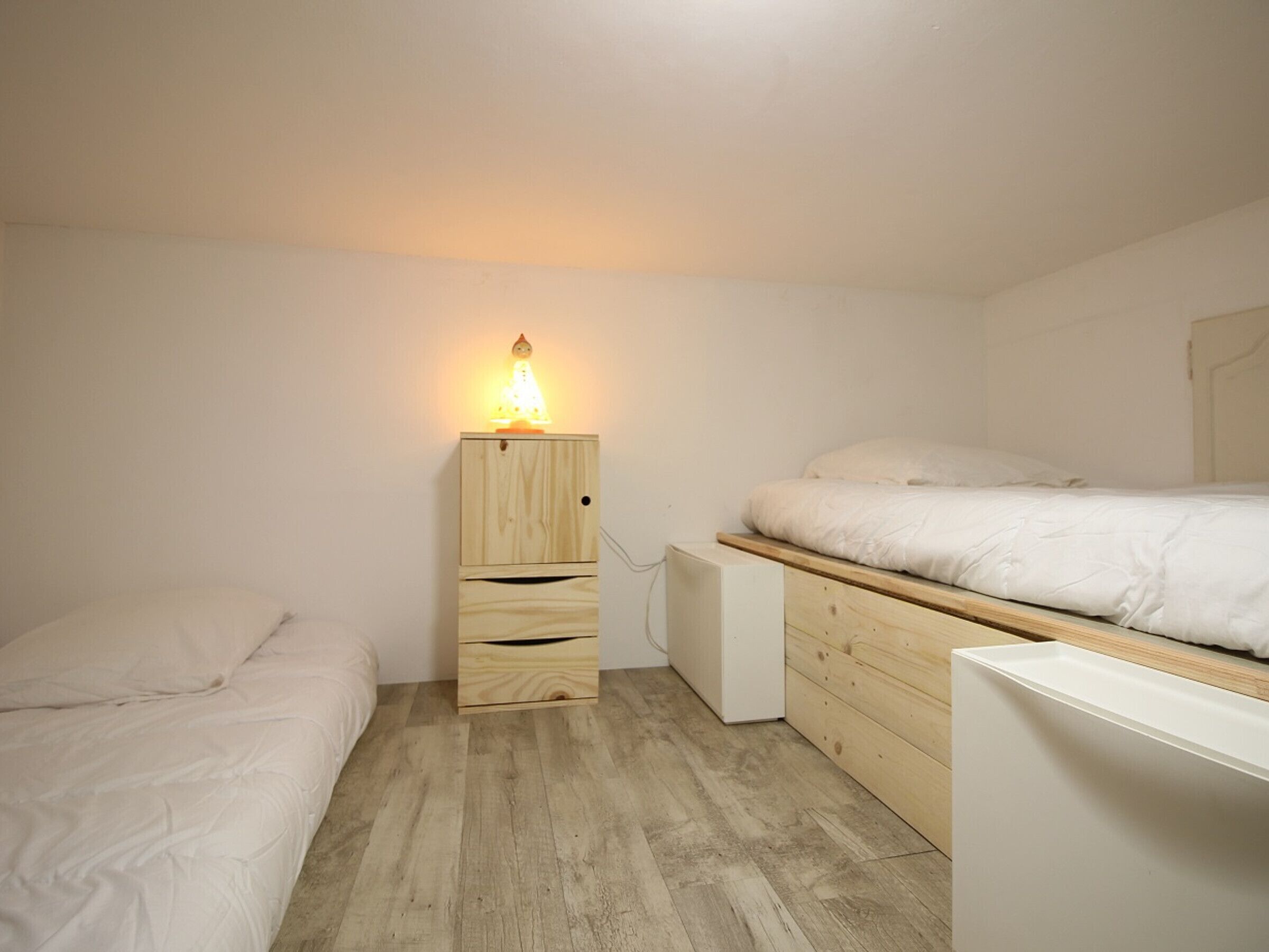 A small, well-lit bedroom with a wooden nightstand, a bedside table, and a bed with white bedding.