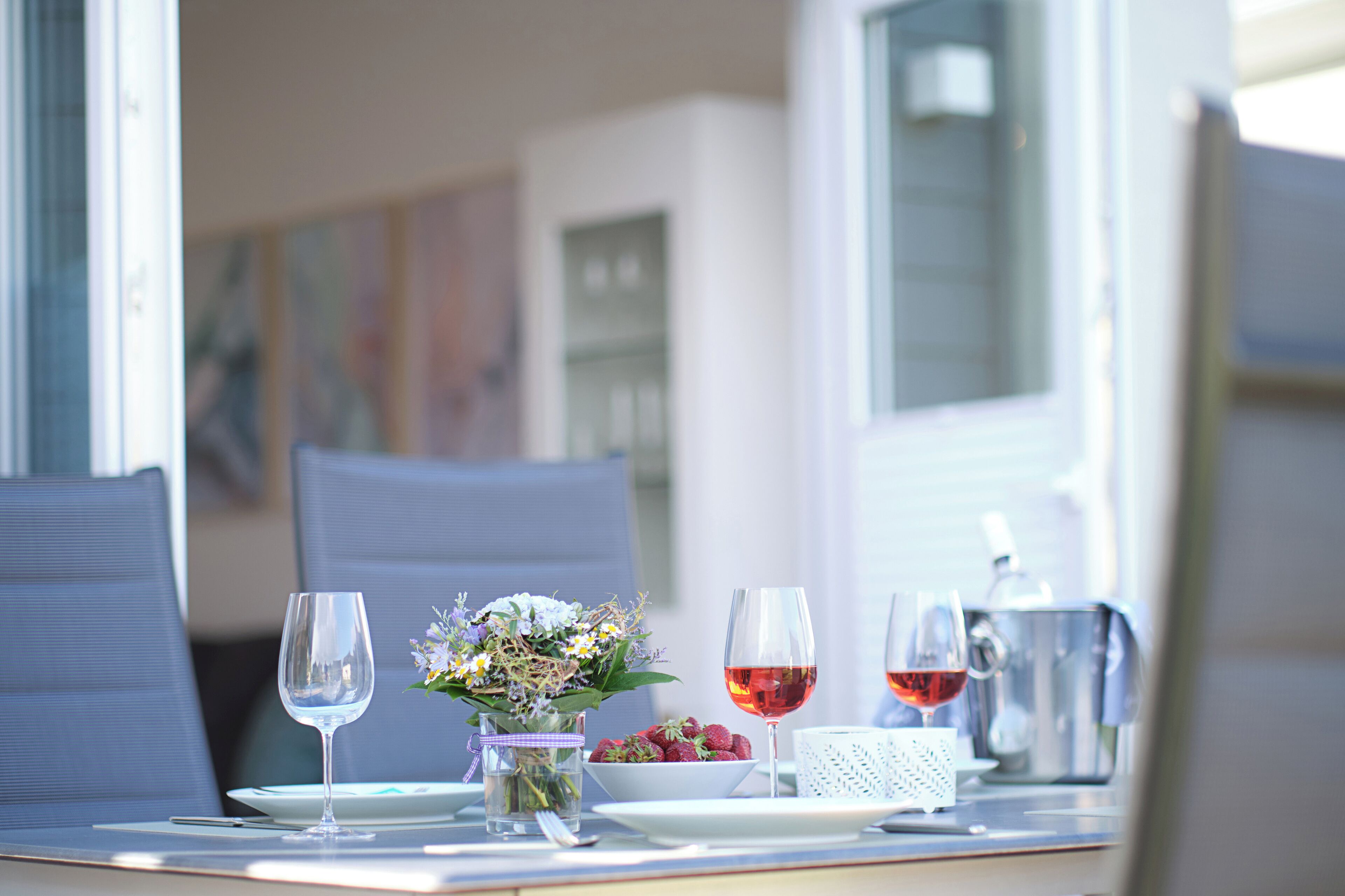 Une table dressée avec un verre d’eau, un bol de fraises, un verre de vin rouge, une cafetière et un vase rempli de fleurs.