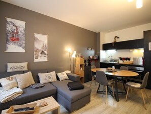 Un salon moderne avec un canapé gris, une table basse en bois et un coin repas comprenant une table ronde et des chaises. On y trouve également une cuisine avec des meubles noirs et un plan de travail blanc.