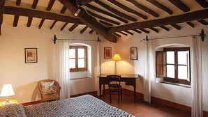 A bedroom with a bed, a desk, and two chairs. There are two windows with curtains, a lamp on the desk, and framed pictures on the walls. - CORZANELLO - Charming farmhouse in the Chianti with  swimming pool use (Calzaiolo)