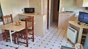 A compact kitchenette with a microwave, a small stove, and a television. There is a dining table with chairs and a cup on it. The room has a tiled floor and wooden cabinets. - Camping Rianxo (Rianxo)