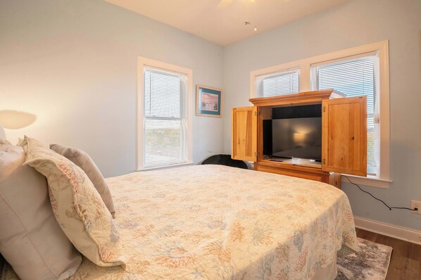 A bedroom with a bed, a wooden cabinet with a television, and two windows with blinds.