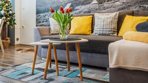 Un salon avec un canapé, une petite table ronde ornée d’un vase de tulipes rouges, et une fresque murale représentant un paysage de montagne.