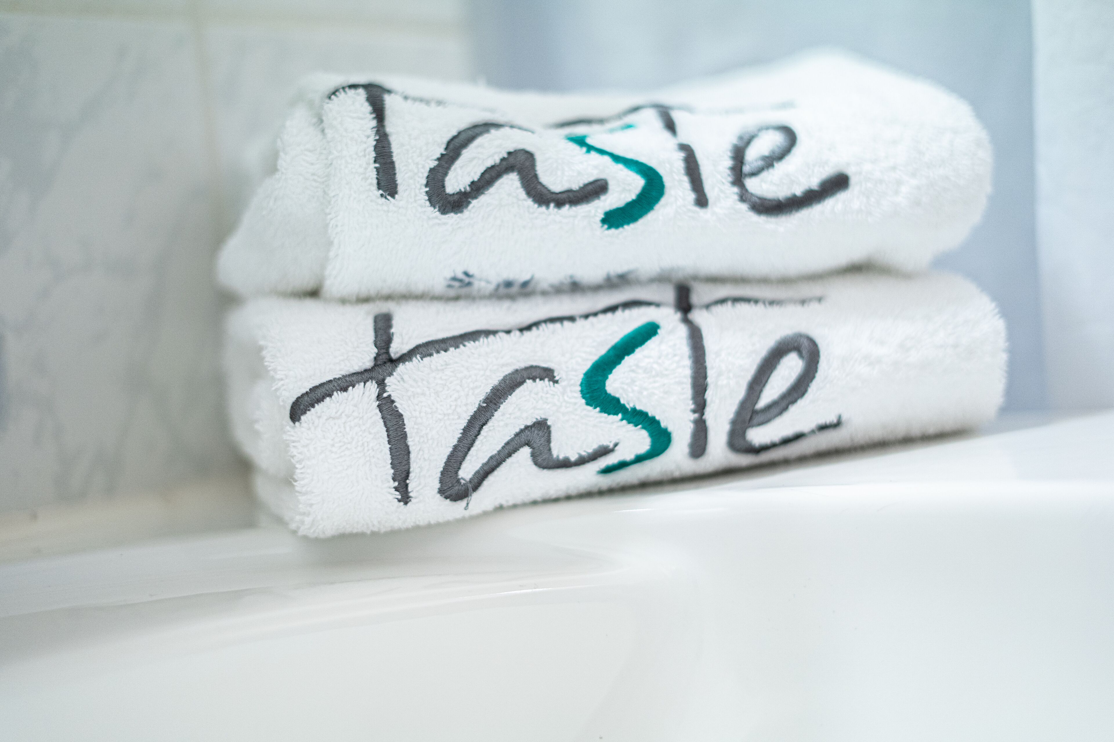 Two white towels stacked on a white surface with the word "Taste" written in black and teal ink.