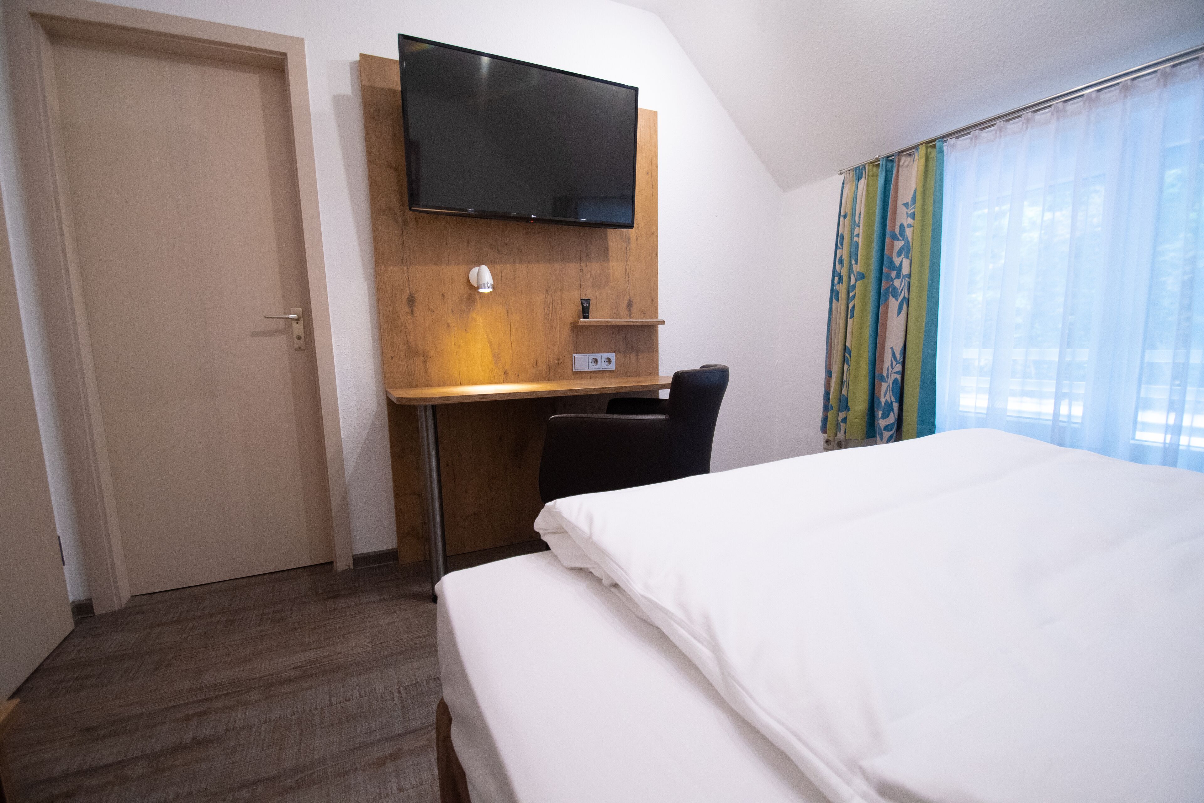 Ein Hotelzimmer mit einem Bett, einem Schreibtisch mit Stuhl, einem an der Wand montierten Fernseher und einem Fenster mit Vorhängen.