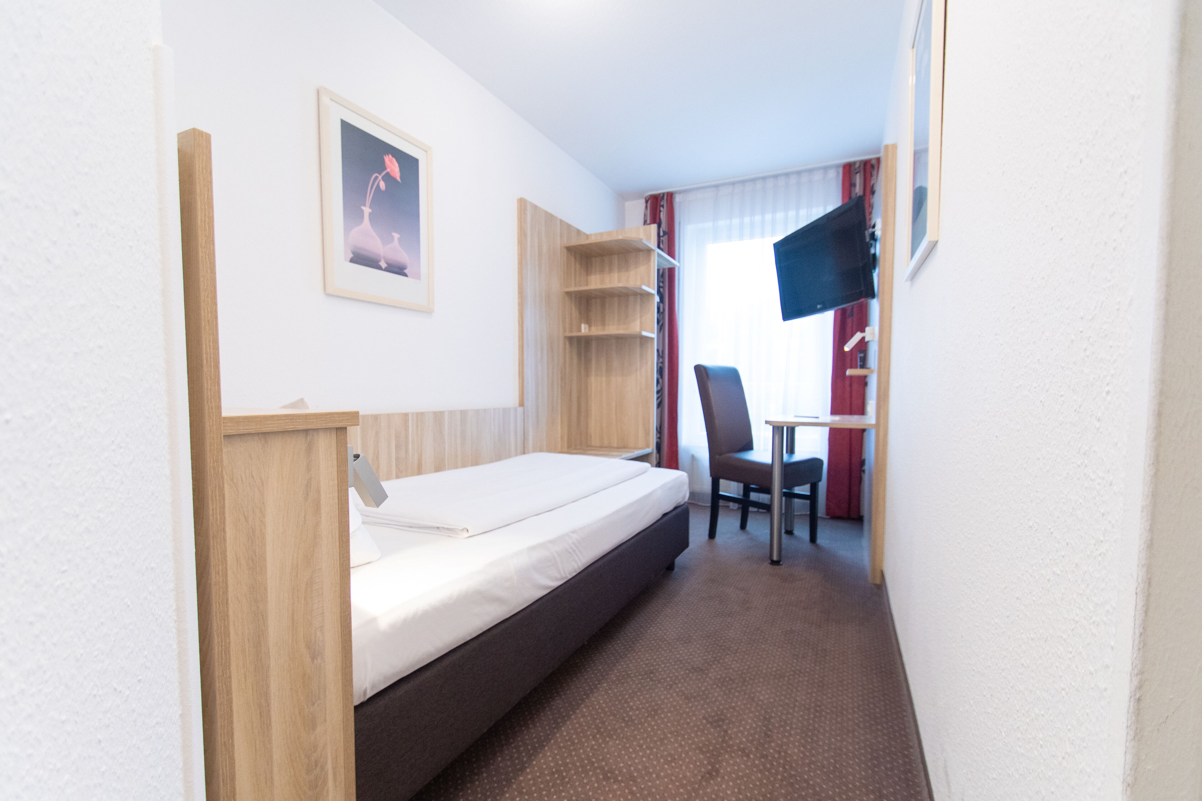 A compact hotel room with a single bed, a small desk, a chair, and a TV.