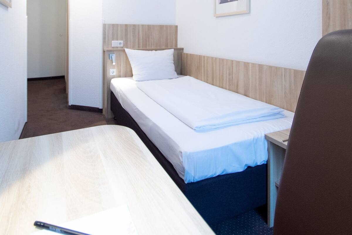 A small, neatly arranged hotel room with a single bed, a desk, and a chair. There is a framed picture on the wall and a door leading to another room.