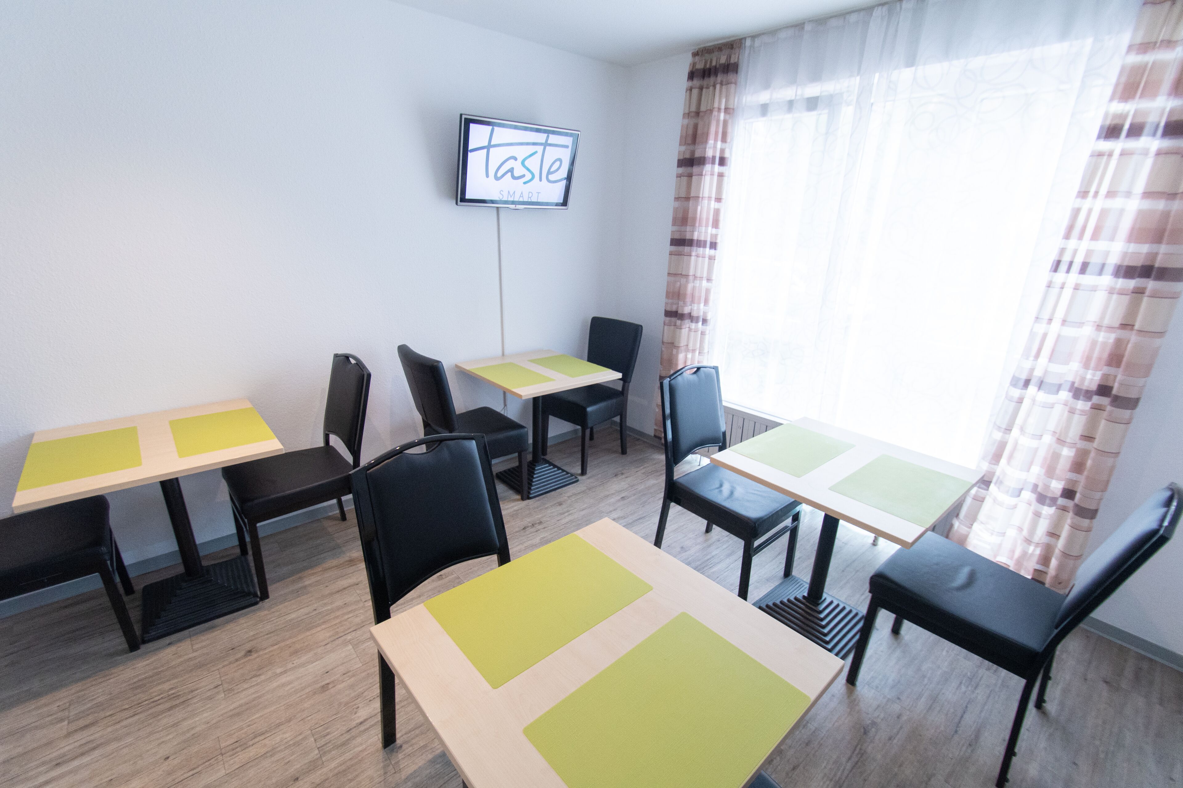 A dining area with multiple tables and chairs, a television mounted on the wall, and a window with curtains.