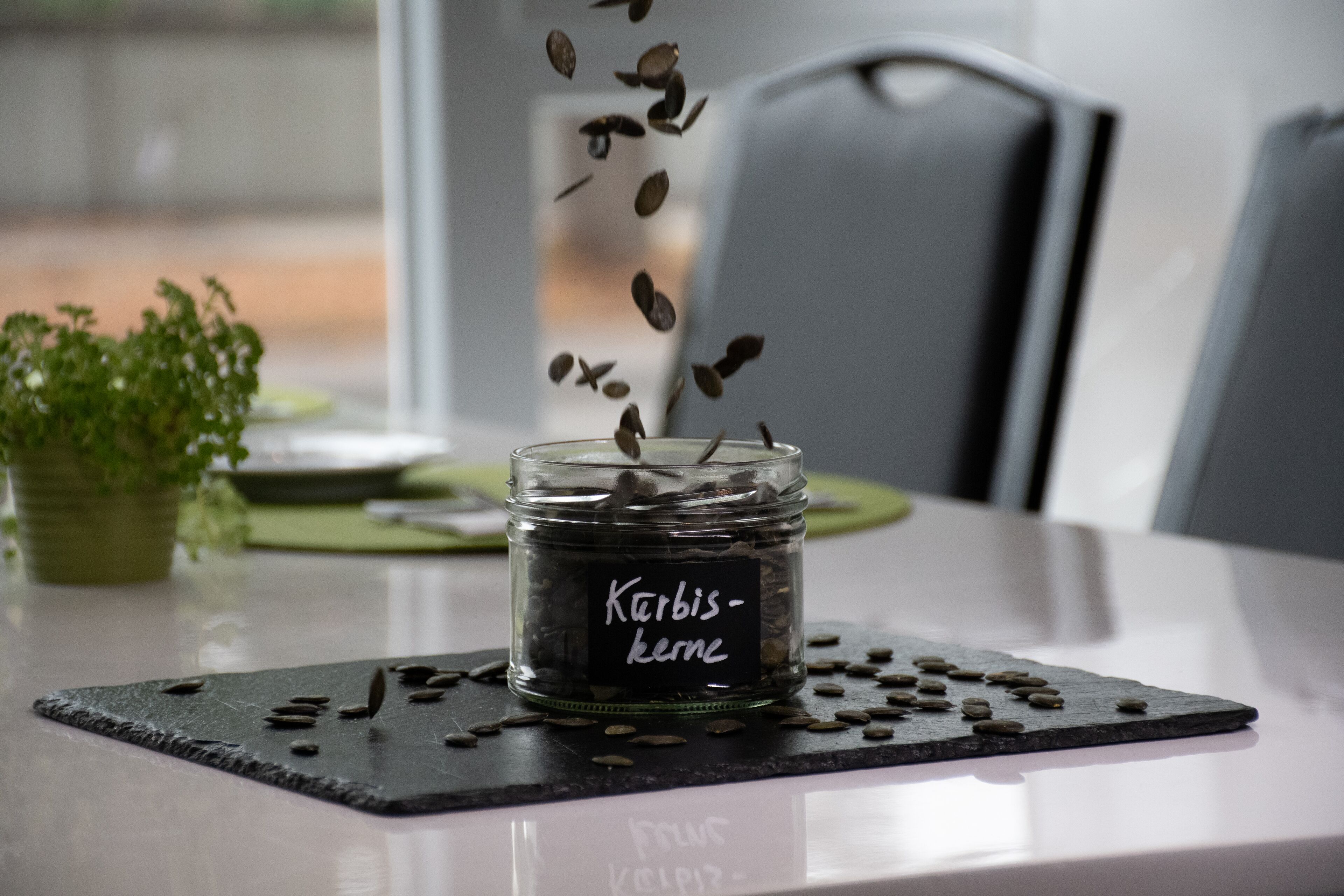 Un frasco de vidrio etiquetado “Karbis-kerne” sobre un posavasos de pizarra negra, con semillas esparcidas.