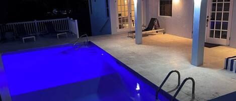 A swimming pool with a blue light, a covered patio area with chairs, and a white fence.