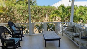 A covered outdoor seating area with wicker chairs and a sofa, a small table, and a view of dense greenery and a building in the background. - Villa - Sleeps 6 - Pool - Parking - Garden (Jolly Harbour)