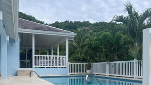 A swimming pool with a white fence, a covered seating area, and lush greenery in the background. - Villa - Sleeps 6 - Pool - Parking - Garden (Jolly Harbour)