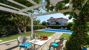 A covered outdoor seating area with wicker furniture, a pool, and a large house in the background. - Stylish sea view studio Artemis, Rhodes-Ialysos (Ialisos)