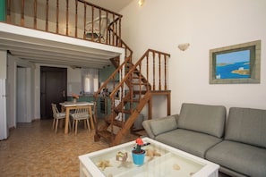A living room with a sofa, a glass coffee table, a dining table with chairs, a staircase, and a painting on the wall.