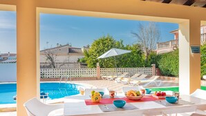 Zona junto a la piscina con una mesa lista para el desayuno, sillas de descanso y vistas a la piscina y edificios circundantes.