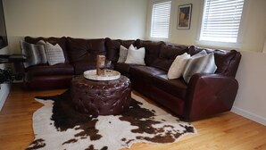 Un salon avec un canapé d’angle en cuir marron, un pouf rond et un tapis en peau de vache.