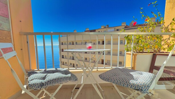Een balkon met twee gestoffeerde stoelen en een kleine tafel, met uitzicht op een kustgebied met gebouwen en een helderblauwe lucht.