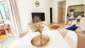 A modern living room with a white sofa, a dining table with a decorative plant, and a kitchen area visible through an archway. - Skol 128 - Great 1-Bedroom Apartment with Sea Views (Marbella)