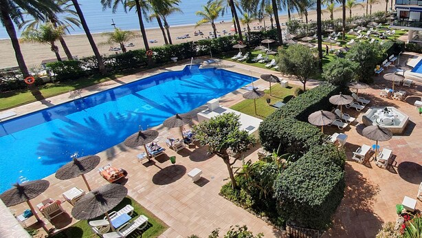 Ein Poolbereich mit Liegestühlen und Sonnenschirmen, umgeben von Palmen und Blick auf den Strand.