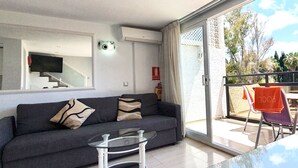 Un salon moderne avec un canapé gris, une table basse en verre et un balcon aménagé avec des sièges d’extérieur.