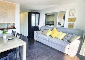 A modern living room with a grey sofa, a dining table with chairs, and a kitchen visible through an open door.
