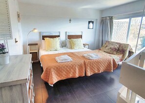 A double bed with an orange quilt, two bedside tables, a nightstand, a window with curtains, and a view of trees.