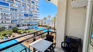 A balcony with a pool, chairs, and a table overlooking a hotel building and beach.