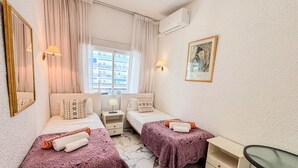 A hotel room with two beds, a small table, a chair, a window with curtains, an air conditioning unit, and a framed picture on the wall.