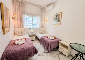 Ein Hotelzimmer mit zwei Betten, einem kleinen Tisch, einem Sessel, einem Fenster mit Vorhängen, einer Klimaanlage und einem gerahmten Bild an der Wand.