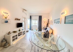 A modern living room with a glass dining table, a sofa, a TV, and artwork on the walls.