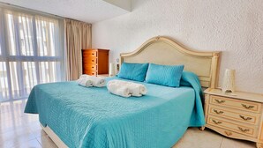 Un dormitori amb un llit gran, una tauleta de nit, una petarrera i una finestra amb cortines.