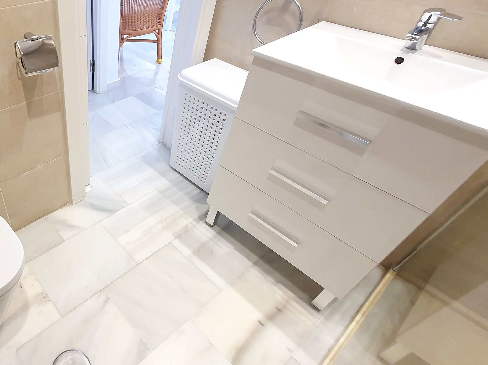 A modern bathroom with a white sink, a toilet, and a wicker chair in the background.