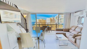 Ein modernes Wohnzimmer mit einem Glastisch, weißen Stühlen, einem beigen Sofa und Blick auf Gebäude durch große Fenster.