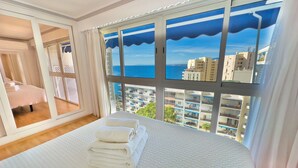 Ein Hotelzimmer mit einem großen Bett, Meerblick und Blick auf Gebäude, sowie ein Balkon mit weißer Brüstung.