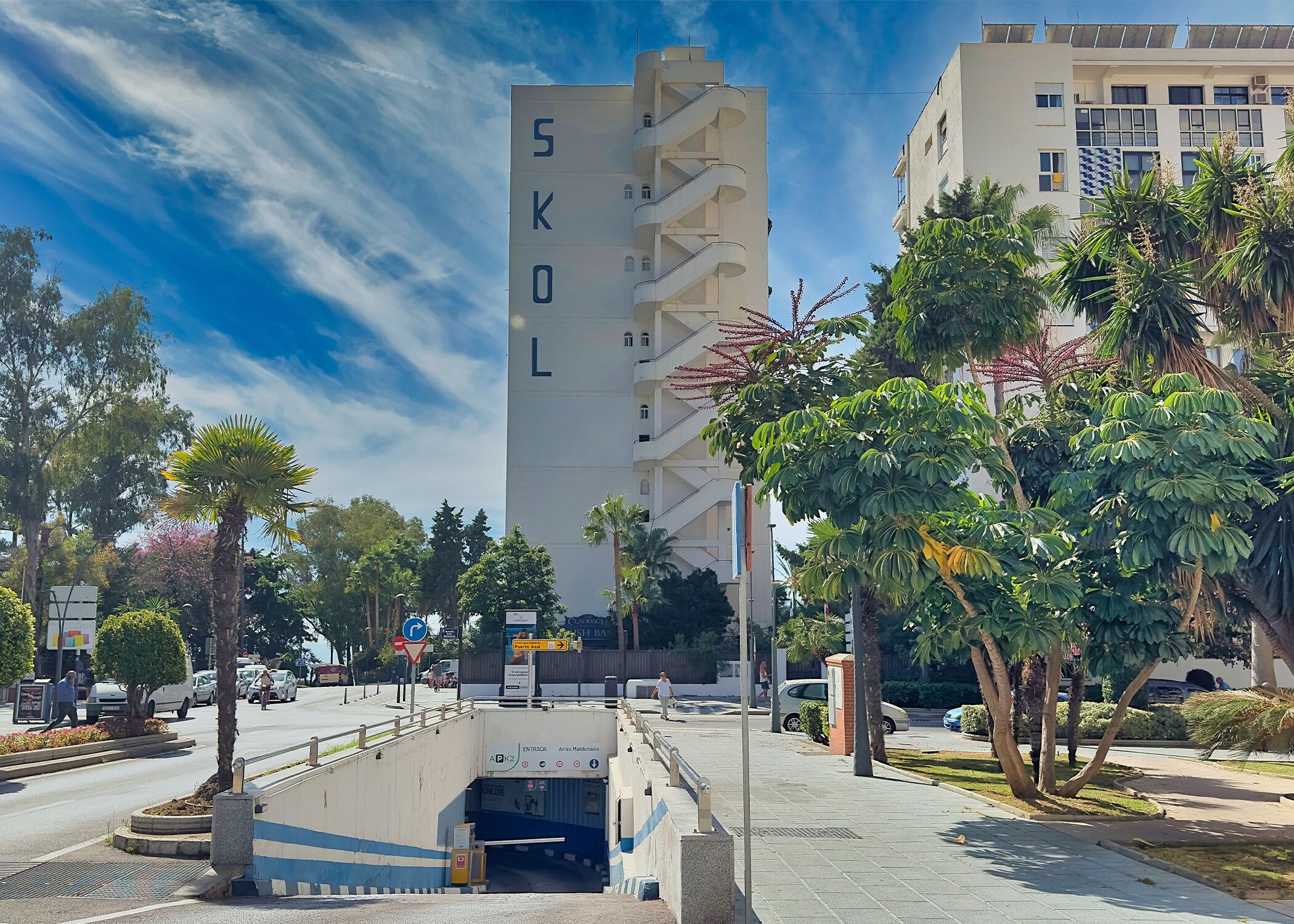 A tall building with 'SKL' on its facade, surrounded by palm trees and a pedestrian walkway.