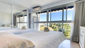 Ein Hotelzimmer mit zwei Betten, großen Fenstern mit Blick auf Bäume und einer Klimaanlage an der Wand.