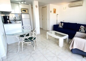 A compact living space with a kitchenette, a dining area with a glass table and white chairs, a coffee table, and a blue sofa with patterned cushions.
