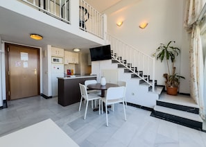 A spacious living area with a dining table, white chairs, a kitchen with a built-in island, a staircase with a white railing, and a potted plant.