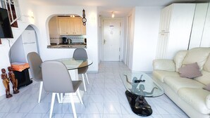 Una sala de estar moderna con una mesa de centro de vidrio, un sofá y un comedor con sillas, con una pequeña cocina al fondo.