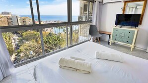 Habitación de hotel con un ventanal grande que ofrece vista a la ciudad, una cama con ropa blanca, un televisor en una cómoda y una silla.