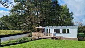Une petite cabane à un seul étage, dotée d’une terrasse, située au bord d’une rivière et entourée d’arbres.
