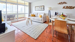 A modern living room with a sofa, dining table, and chairs. There is a large window with a view of buildings.