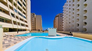 A swimming pool with a geometric pool edge design, surrounded by high-rise buildings with balconies and a clear blue sky.